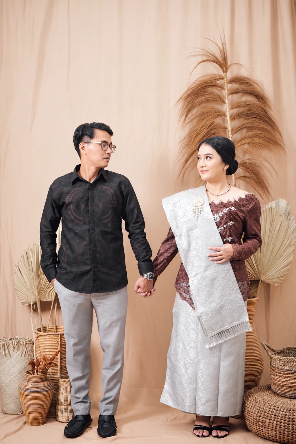 Ryan berkemeja hitam dan Santi berkebaya bergandengan tangan di depan latar peach